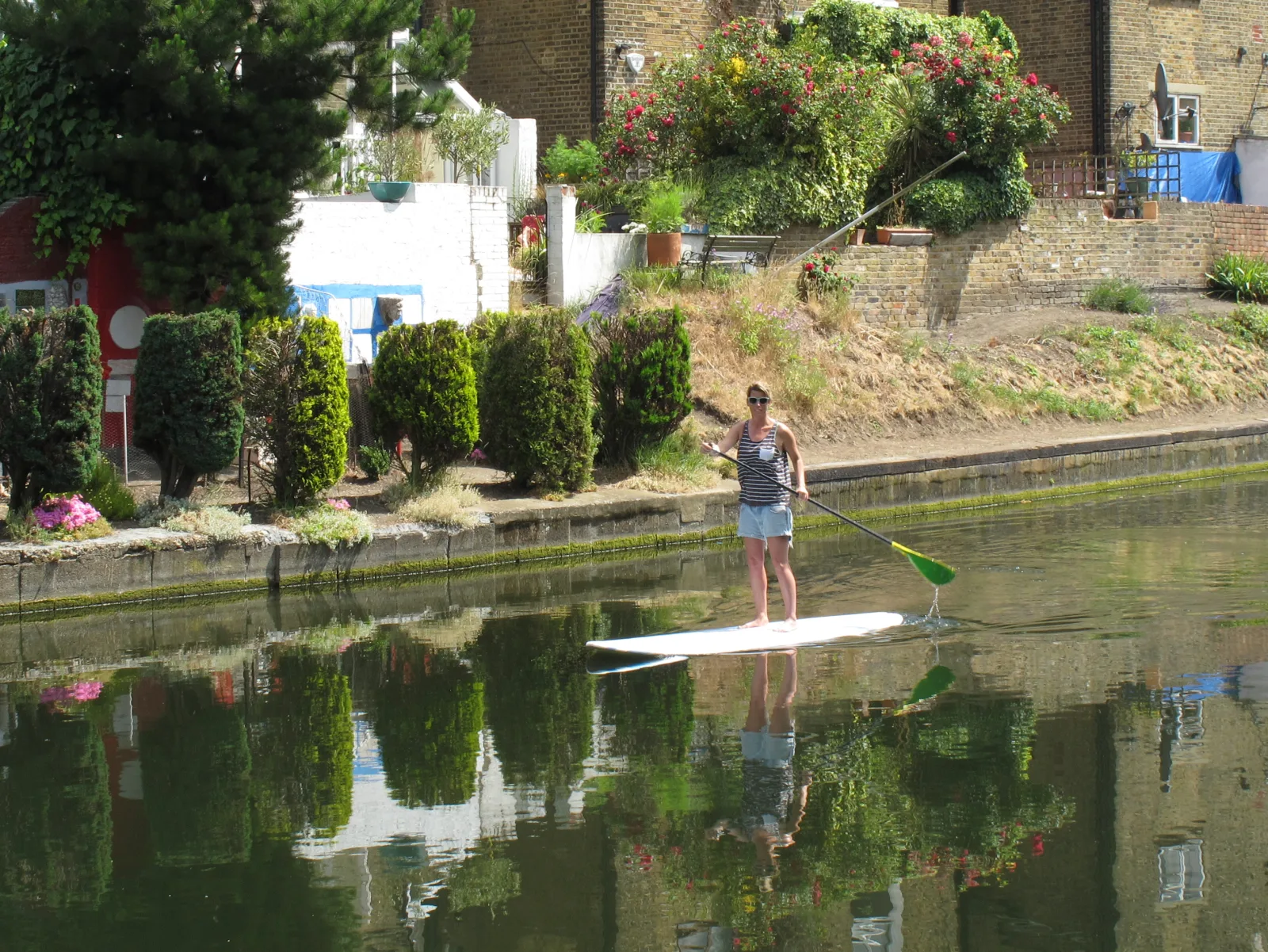 Vrouw op een SUP paddleboard op een rustig kanaal met groene oever en weerspiegeling van tuinen in het water